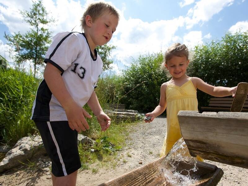 Zwei Kinder spielen an einem Wasserspielplatz im Freien, umgeben von Grün und unter blauem Himmel.