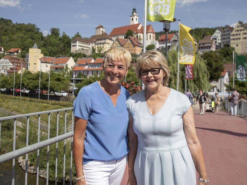 Zwei Frauen stehen lächelnd auf einer Brücke mit einer Stadt im Hintergrund, wo bunte Fahnen wehen und Gebäude an einem bewaldeten Hügel zu sehen sind.