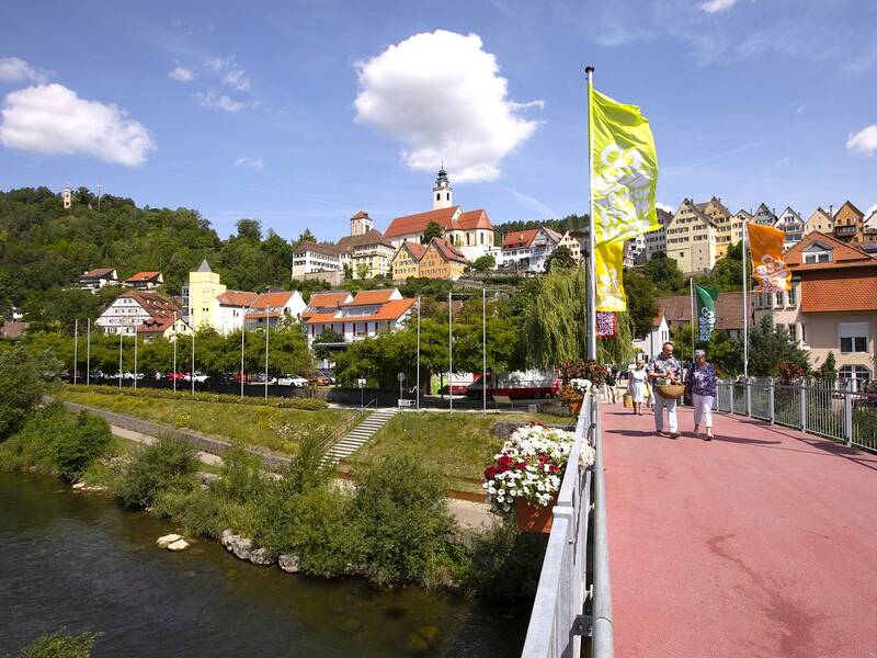 Eine malerische Stadtansicht zeigt eine Brücke mit Personen, umgeben von traditionellen Häusern und üppiger Natur unter einem blauen Himmel mit ein paar Wolken.