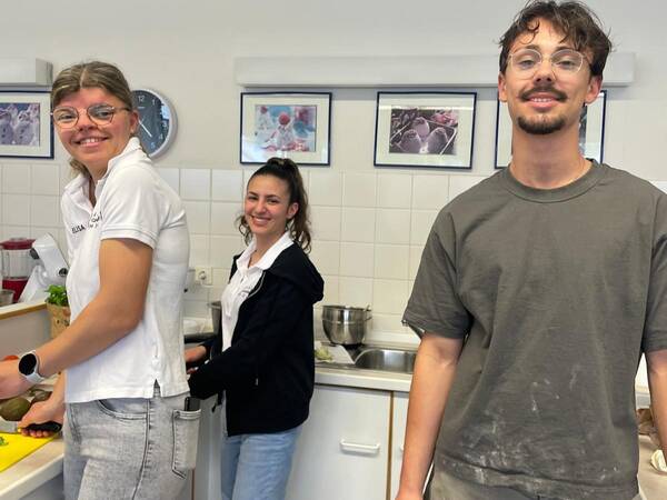 Drei Personen in einer Küche während eines Kochworkshops. Eine Person schneidet Gemüse, eine andere steht am Waschbecken. Die dritte Person posiert in der Nähe.