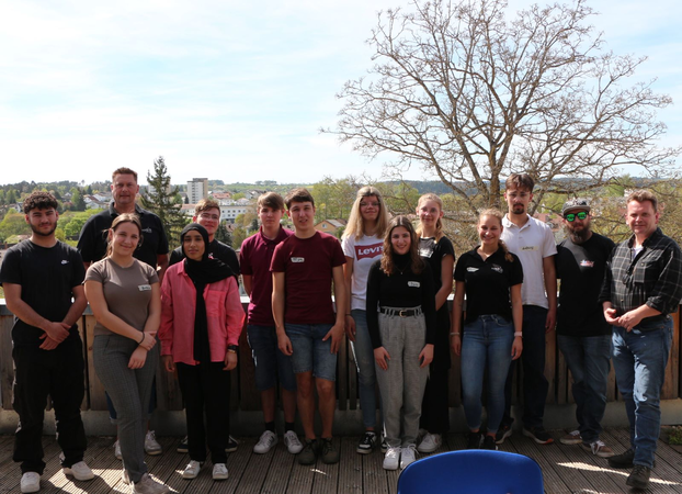 Eine Gruppe von 14 Personen steht auf einer Terrasse, im Hintergrund sind Bäume und Gebäude zu sehen.