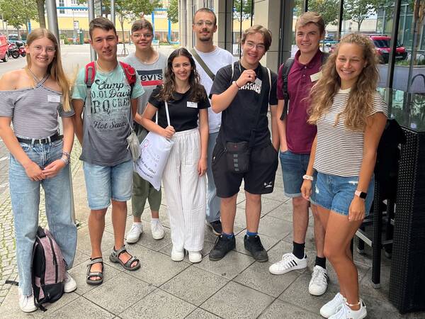 Eine Gruppe von acht jungen Menschen steht lächelnd auf einem Gehweg in der Nähe eines Gebäudes.