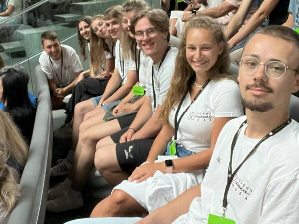 Eine Gruppe von jungen Erwachsenen sitzt in einer Reihe auf Tribünenplätzen und lächelt in die Kamera.