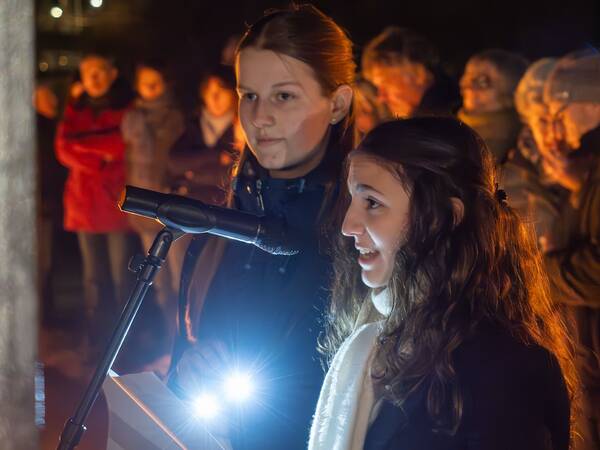 Zwei junge Frauen stehen im Dunkeln vor einem Mikrofon, während hinter ihnen eine Gruppe von Menschen unscharf zu erkennen ist.