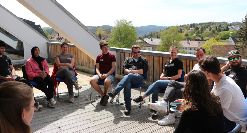 Eine Gruppe von Menschen sitzt in einem Kreis auf einer Terrasse und unterhält sich, während die Sonne scheint und im Hintergrund eine hügelige Landschaft zu sehen ist.