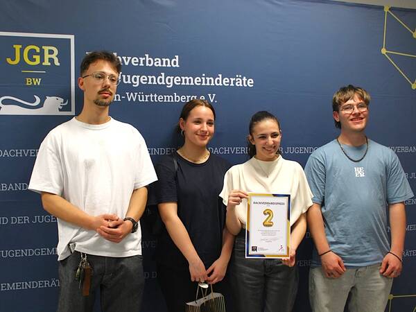 Vier Personen stehen vor einem blauen Hintergrund mit dem Logo 'JGR BW'. Eine Person hält ein Zertifikat mit der Aufschrift '2. Platz'.