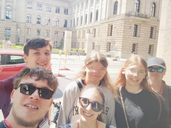 Eine Gruppe von sechs Personen macht bei sonnigem Wetter vor einem eindrucksvollen historischen Gebäude ein Selfie.