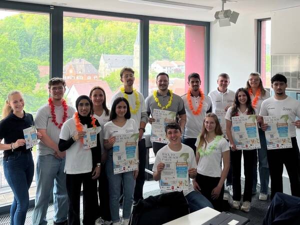Eine Gruppe von Menschen posiert in einem Raum vor großen Fenstern, lächelt und hält farbenfrohe Poster und Flyer, während einige Erinnerungsstücke wie Blumenkränze tragen.