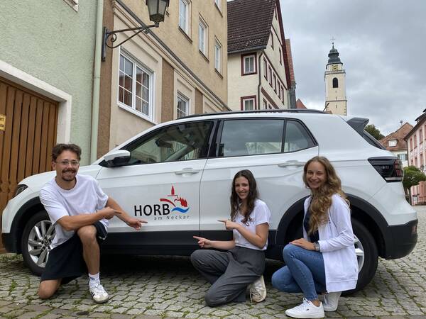 Drei lächelnde Personen zeigen auf ein Auto mit der Aufschrift "Horb am Neckar", das in einer gepflasterten Straße vor historischen Gebäuden und einer Kirchturmspitze im Hintergrund steht.