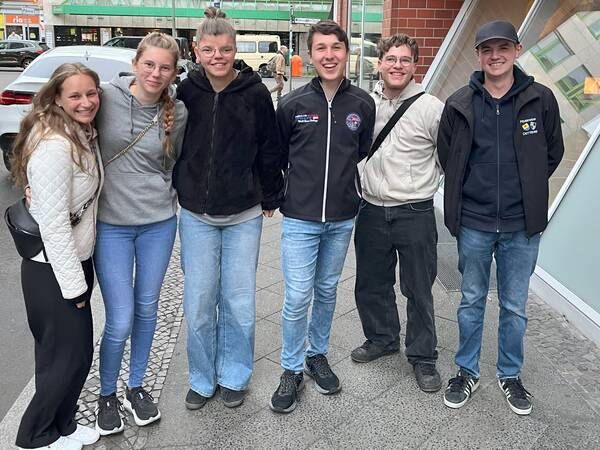 Sechs junge Erwachsene stehen lächelnd nebeneinander auf einem Gehweg vor einem Gebäude.