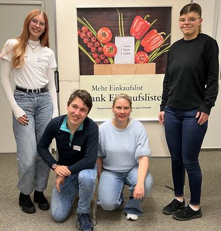 Vier Personen posieren vor einem Poster, das eine Einkaufsliste und Gemüse zeigt.