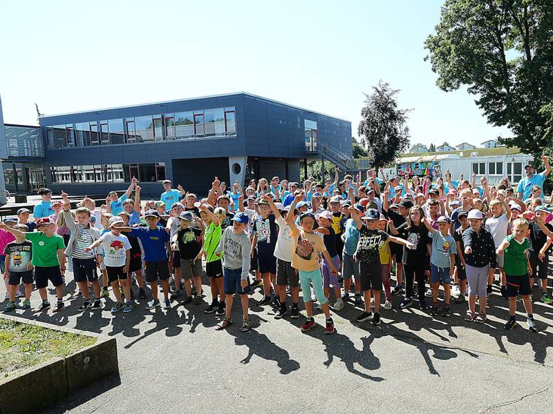 Viele Kinder und Betreuuende auf dem Pausenhof im Schulzentrum, die tanzend die Hände in die Luft halten.