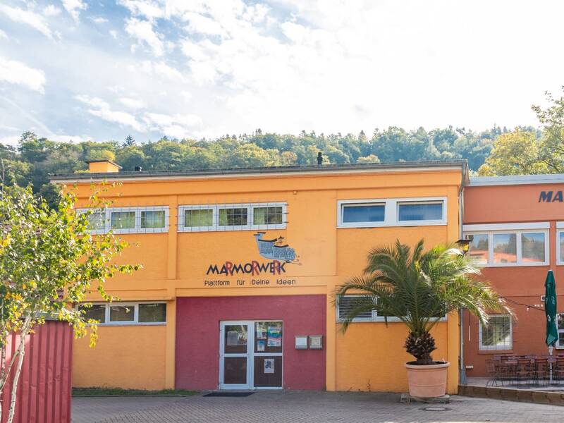 Gelb-orangefarbenes Gebäude mit dem Schriftzug 'MARMORWERK' und einer Palme vor dem Eingang.