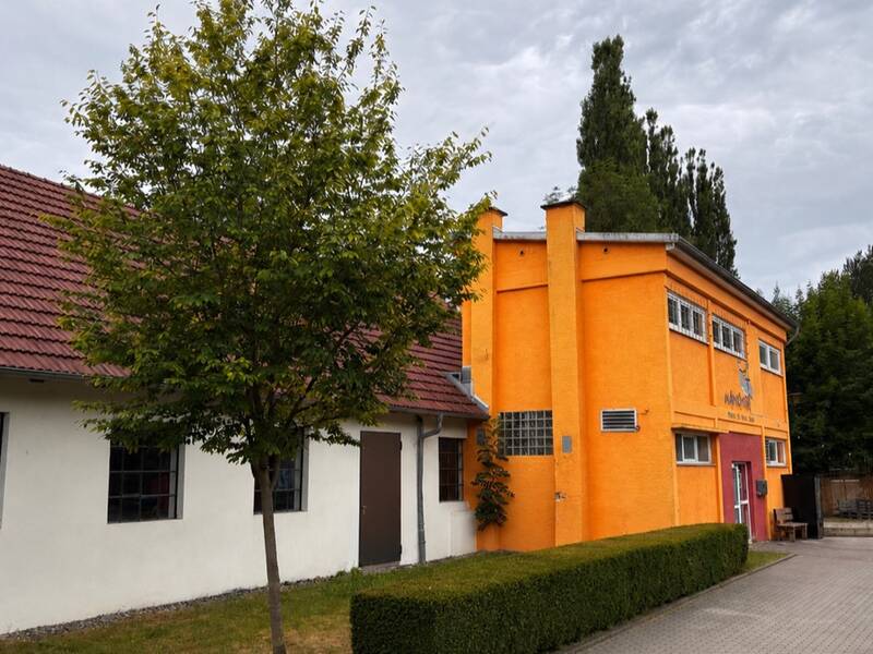 Das Bild zeigt ein Gebäude mit einem orangefarbenen Abschnitt und einem weißen Abschnitt, umgeben von Bäumen und einem gepflasterten Weg.