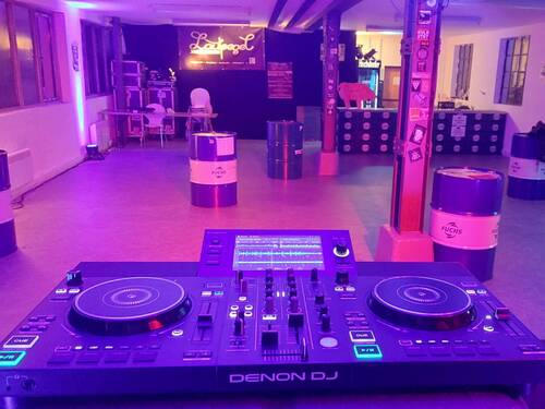 Ein mit lila Licht beleuchteter Raum enthält ein DJ-Pult im Vordergrund und mehrere dekorative Fässer im Hintergrund.