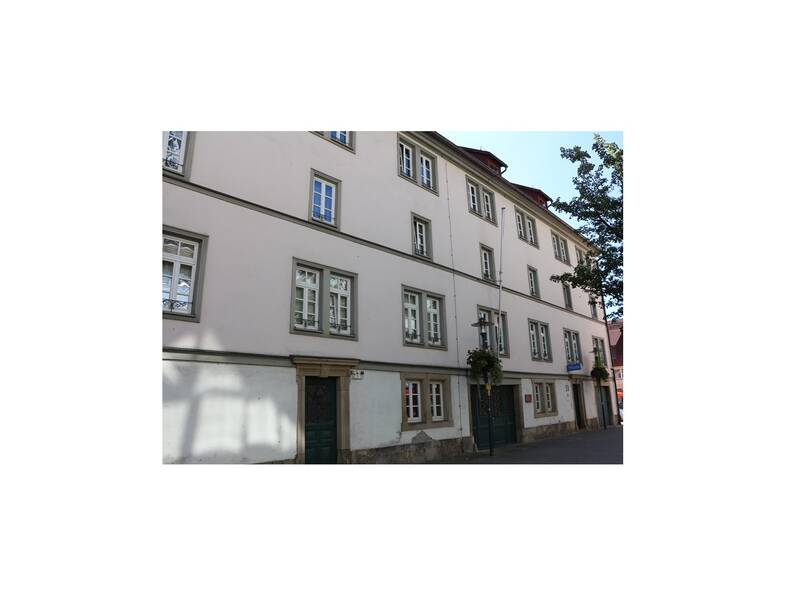 Das Bild zeigt ein großes, mehrstöckiges Gebäude mit weißen Wänden, zahlreichen Fenstern und einem Eingang mit grüner Tür in einer städtischen Umgebung.