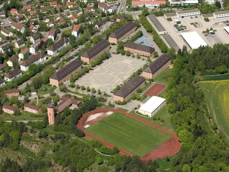 Luftbild einer Stadt mit mehreren langen Gebäuden in einem rechteckigen Hof, umgeben von Wohnhäusern, Industriegebäuden, Grünflächen und einem Sportplatz.