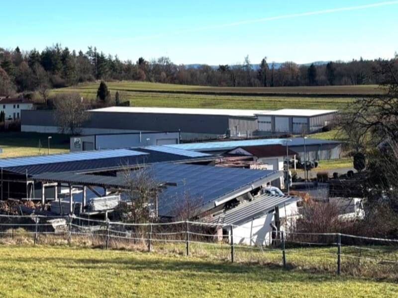 Eine ländliche Landschaft mit Wiesen im Vordergrund, mehreren Industriegebäuden in der Mitte und einem Wald am Horizont unter einem klaren blauen Himmel.
