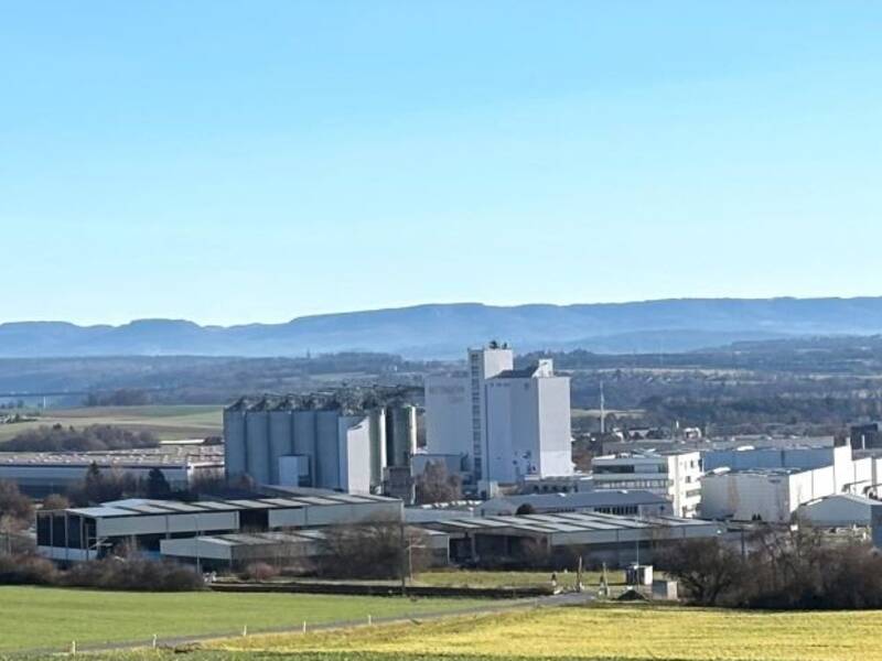 Weitläufige Landschaft mit Feldern im Vordergrund, einer Industrieanlage in der Mitte vom Industriegebiet Heiligenfeld und einem bewaldeten Gebirgszug im Hintergrund unter blauem Himmel.