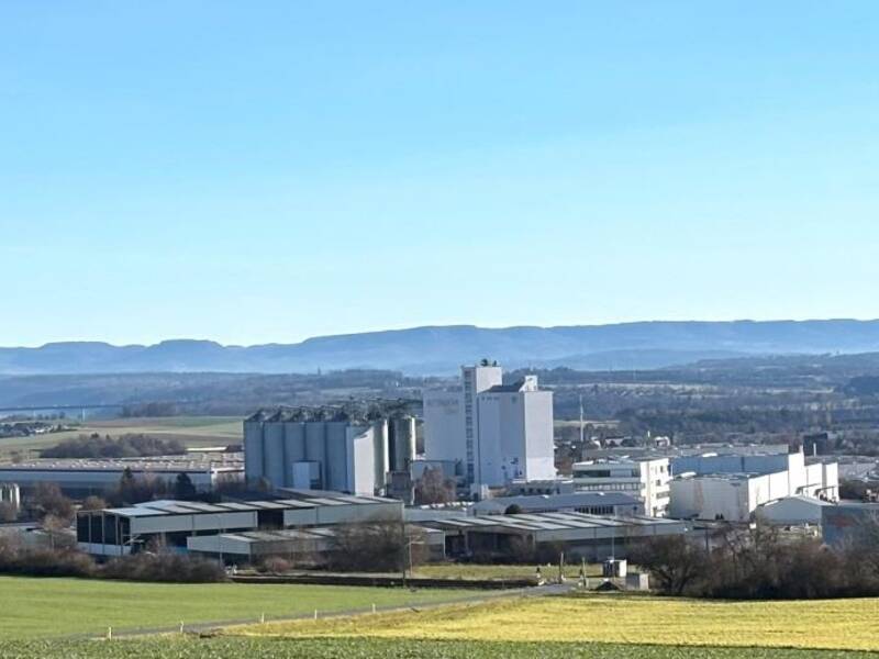 Weitläufige Landschaft mit Feldern im Vordergrund, einer Industrieanlage in der Mitte vom Industriegebiet Heiligenfeld und einem bewaldeten Gebirgszug im Hintergrund unter blauem Himmel.