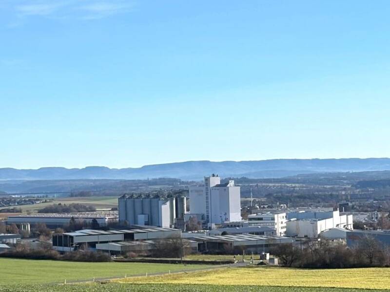 Weitläufige Landschaft mit Feldern im Vordergrund, einer Industrieanlage in der Mitte vom Industriegebiet Heiligenfeld und einem bewaldeten Gebirgszug im Hintergrund unter blauem Himmel.