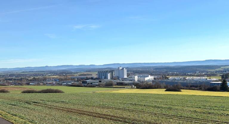 Weitläufige Landschaft mit Feldern im Vordergrund, einer Industrieanlage in der Mitte vom Industriegebiet Heiligenfeld und einem bewaldeten Gebirgszug im Hintergrund unter blauem Himmel.