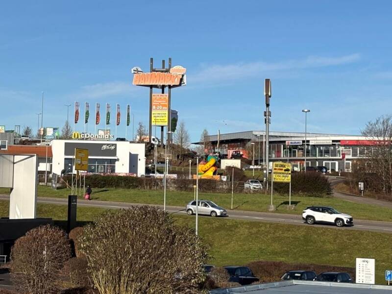 Die Szene zeigt das Gewerbegebiet Hohenberg mit einem Fast-Food-Restaurant und einem Autohaus im Vordergrund, umgeben von geparkten Autos und einer Straße, auf der ein weißes Auto fährt.