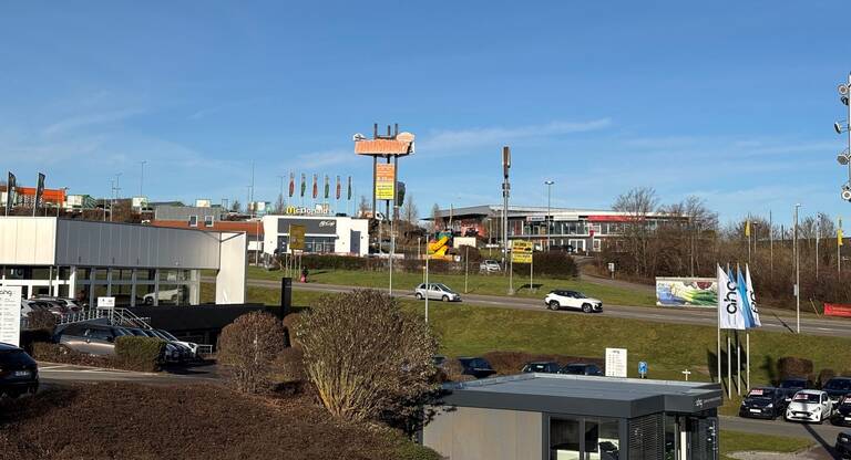 Die Szene zeigt das Gewerbegebiet Hohenberg mit einem Fast-Food-Restaurant und einem Autohaus im Vordergrund, umgeben von geparkten Autos und einer Straße, auf der ein weißes Auto fährt.