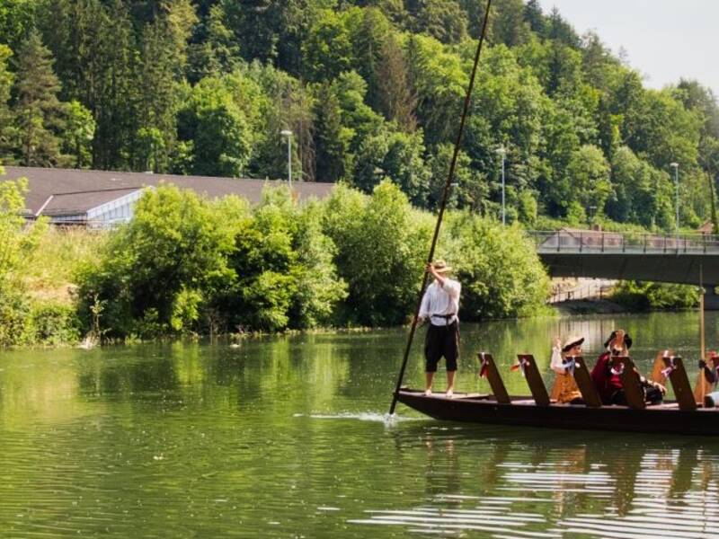 Ein Flachboot mit mehreren sitzenden Personen fährt auf einem Fluss, während am Ufer weitere Menschen in sommerlicher Kleidung stehen und zuschauen, umgeben von grüner Vegetation und Hügeln im Hintergrund.
