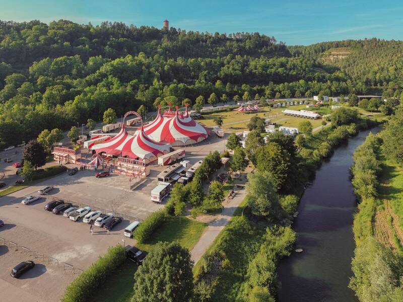 Eine Luftaufnahme zeigt ein großes rot-weiß gestreiftes Zirkuszelt inmitten einer von Bäumen umgebenen Landschaft, mit einem Parkplatz und mehreren Wohnmobilen daneben sowie einem Fluss, der rechterhand fließt.