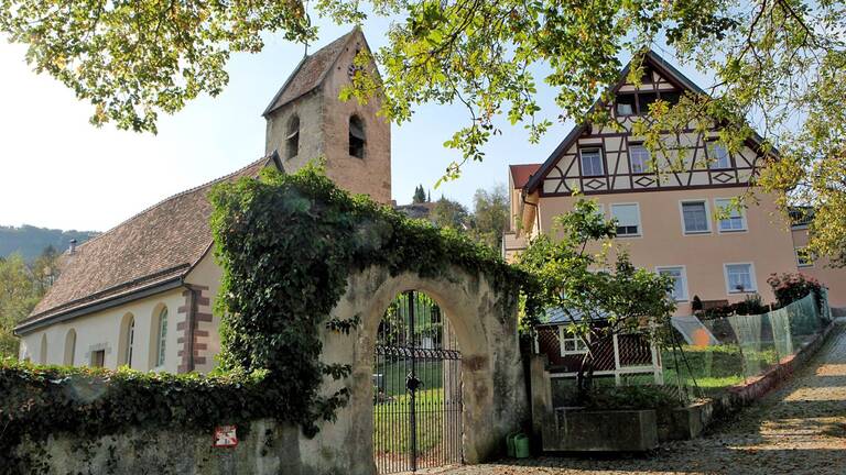 Eine kleine Kirche mit einem Glockenturm und ein traditionelles Fachwerkhaus stehen nebeneinander, umgeben von Bäumen und einem gepflasterten Weg.