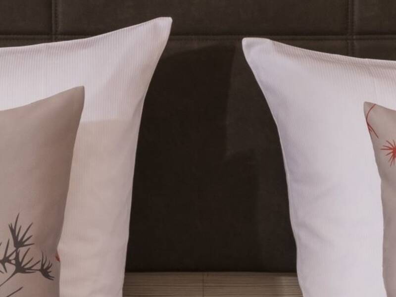 Zwei dekorative Kissen mit floralen Mustern in Rot und Grau liegen nebeneinander auf einem Bett mit einem dunkel gepolsterten Kopfteil.