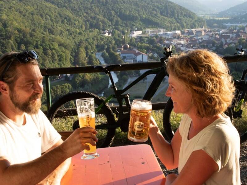 Ein Mann und eine Frau mit einem Bier in der Hand, die an einem Tisch im Biergarten sitzen. Im Hintergrund sieht man den Ausblick auf eine Stadt