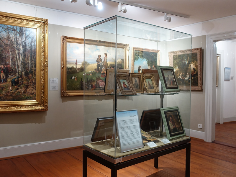 Ein Raum mit einer Glasvitrine, in der Bilder ausgestellt werden. An den Wänden hängen gemälde in goldenen Rähmen.