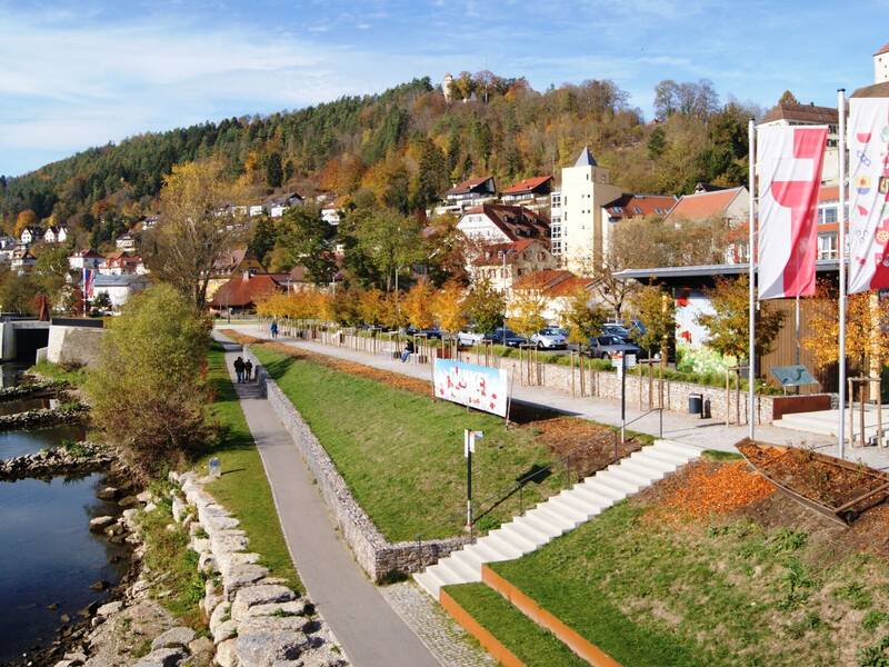 Eine malerische Neckarlandschaft mit einem Uferweg, herbstlich gefärbten Bäumen, mehreren Flaggen und im Hintergrund der Horber Flößerweg an einem bewaldeten Hang.