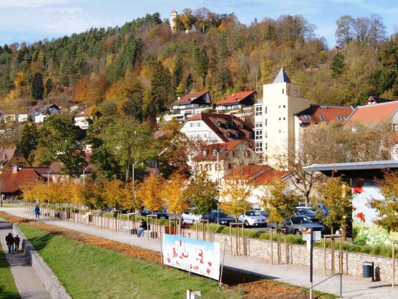 Eine malerische Neckarlandschaft mit einem Uferweg, herbstlich gefärbten Bäumen, mehreren Flaggen und im Hintergrund der Horber Flößerweg an einem bewaldeten Hang.