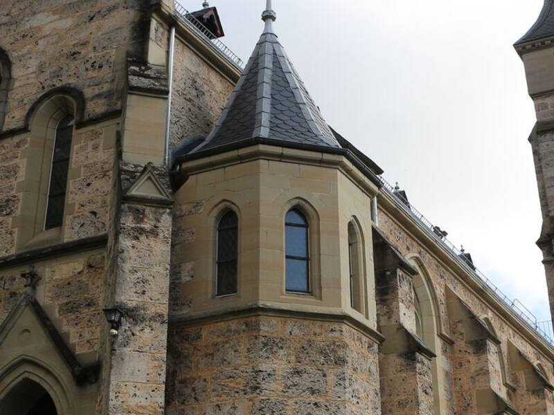 Das Bild zeigt eine große, historische Kirche mit einem hohen Glockenturm und verzierten gotischen Fenstern, umgeben von Pflanzen.
