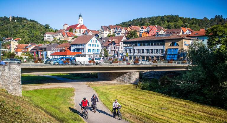 Drei Personen fahren mit Fahrrädern auf dem Neckartalradweg unter einer Brücke entlang, mit der Horber Stadt voller bunter Häuser und einer Kirche im Hintergrund.