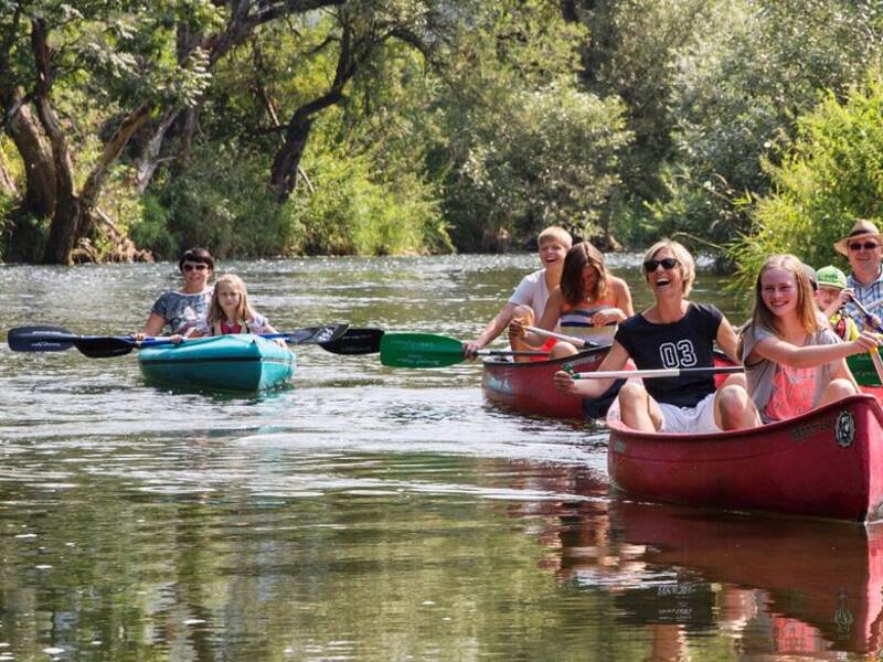 Eine Gruppe von Menschen paddelt in Kanus auf einem von Bäumen gesäumten Fluss.