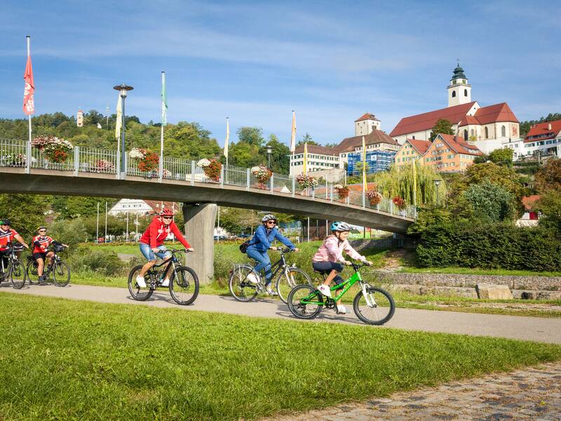 Eine Gruppe von Radfahrern fährt einen Weg entlang, während im Hintergrund eine Brücke mit Blumenschmuck und eine Kirche der Stadt Horb am Neckar zu sehen sind.