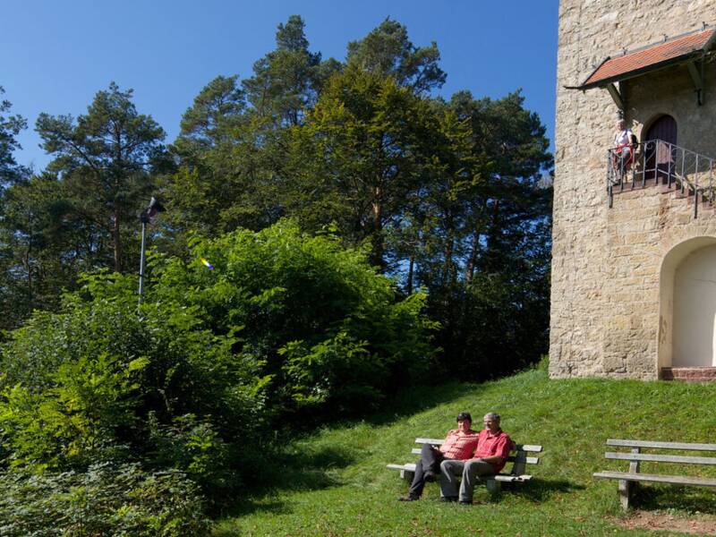Ein alter Steinturm, der Schütteturm, steht in einer grünen Landschaft mit Bäumen und einem blauen Himmel, während zwei Personen auf einer Bank davor sitzen.