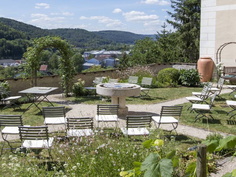 Ein Garten mit Tischen und Stühlen bietet einen weiten Blick auf bewaldete Hügel und einen Fluss, neben einem Gebäude mit grünen Fensterläden.