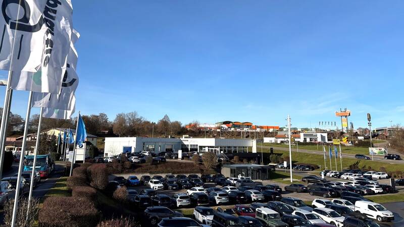 Ein Parkplatz im Gewerbegebiet Hohenberg mit vielen Autos vor einem Gebäude, im Hintergrund sind Fahnen und ein Hügel mit verschiedenen Gebäuden und einer Werbetafel zu sehen.