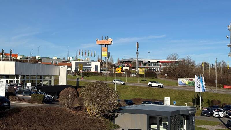 Die Szene zeigt das Gewerbegebiet Hohenberg mit einem Fast-Food-Restaurant und einem Autohaus im Vordergrund, umgeben von geparkten Autos und einer Straße, auf der ein weißes Auto fährt.