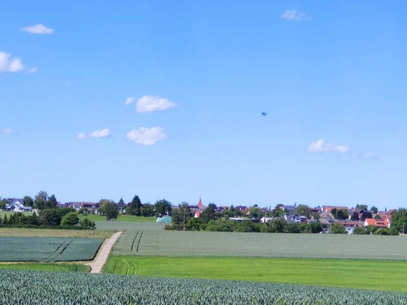 Blick auf einen Ort mit roten Dächern hinter grünen Feldern unter blauem Himmel mit vereinzelten Wolken.