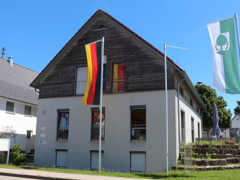 Ein zweistöckiges Gebäude mit der Nummer 21 ist zwischen zwei Fahnen positioniert, einer deutschen Flagge und dem Ahldorfer Stadtwappen, umgeben von einem Vorgarten und einer Straße bei klarem Himmel.