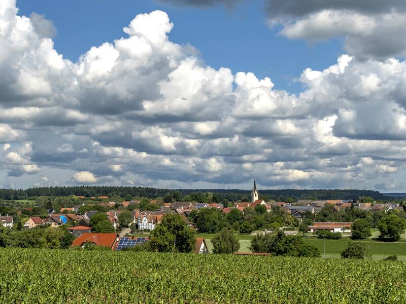 Eine ländliche Landschaft mit einem Dorf im Hintergrund, einer Kirche mit einem hohen Turm und weiten Feldern im Vordergrund unter einem bewölkten Himmel.