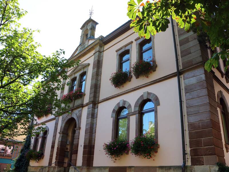 Ein historisches Gebäude mit blassrosa Wänden, großen Fenstern mit Blumenkästen darunter und einem kleinen Turm auf dem Dach, umgeben von grünem Laub.
