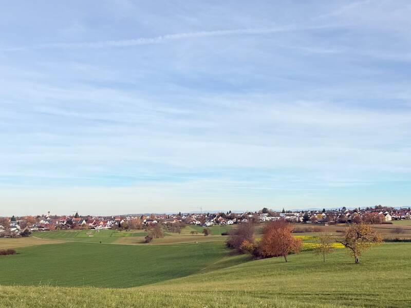 Eine malerische Dorflandschaft mit roten Dächern und einem Kirchturm unter einem blauen Himmel mit weißen Wolken.