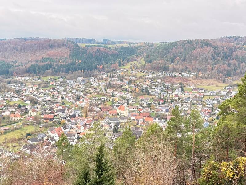 Die Luftaufnahme zeigt ein Dorf mit einer zentral gelegenen Kirche und umliegenden Häusern in einer grünen, hügeligen Landschaft.